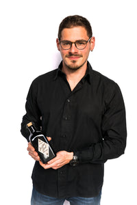 Junger Mann mit Brille, kurzen braunen Haaren, Bart , Brille und schwarzem Hemd lacht in die Kamera und hält eine schwarze Flasche Buddas Hand Gin